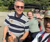 Three people are smiling for a selfie with the person in the foreground giving a thumbs up and the two in the background seated on a park bench waving at the camera