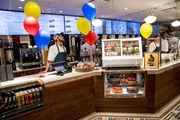 A neatly organized deli counter featuring a staff member, various foods on display, festive balloons, and a menu board in the background.