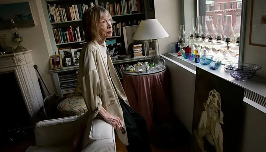 A woman stands contemplatively in a cozy, book-filled room with an elaborate collection of glassware displayed on a windowsill.