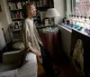 A woman stands contemplatively in a cozy room filled with books and eclectic decor looking toward a window that reveals an urban view