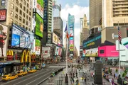 The image captures a bustling Times Square in New York City, dotted with bright digital billboards, yellow taxis, and pedestrians.