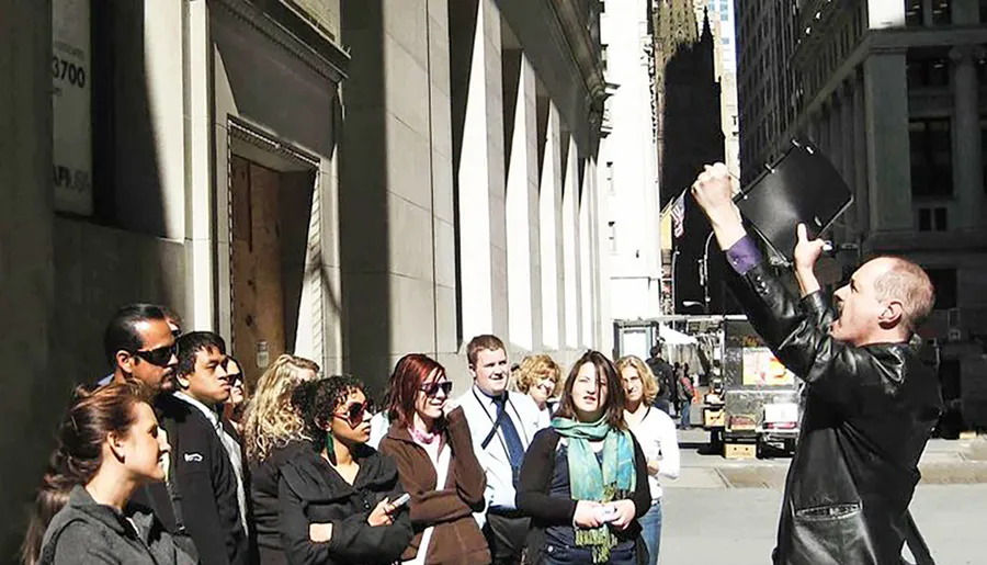 A group of people watches attentively as a man holding a black portfolio looks up, demonstrating something to them on a sunny day in an urban setting.