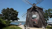 A traditional wooden windmill stands by a clear blue sky with an American flag out front, while people enjoy a sunny day by the water.