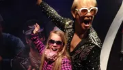 A child and an adult are posing joyfully with raised arms, both wearing large sunglasses and colorful outfits, against a dark backdrop with partial silhouettes of other figures.