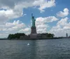 The image shows the Statue of Liberty on its island under a partly cloudy sky as seen from a distance over the water with a buoy visible in the foreground