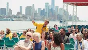 A tour guide in a yellow jacket is speaking into a microphone and pointing something out to a diverse group of attentive passengers on a boat tour with a city skyline in the background.