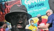 A man in a black hat is taking a selfie with a colorful graffiti wall in the background, while several people, including one wearing a mask, are engaging with each other in the background.