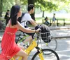 A person in a red dress is riding a yellow bicycle in a park with a helmet in the front basket while there are other people in the background possibly enjoying a sunny day outdoors