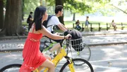 A person in a red dress is riding a yellow bicycle in a park with a helmet in the front basket, while there are other people in the background, possibly enjoying a sunny day outdoors.