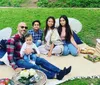 A family enjoys an outdoor picnic on a blanket with food and drinks while smiling and posing for a photo in a grassy park area