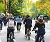 Two people with helmets are smiling for a selfie with a park featuring trees and people in the background suggesting they might be enjoying an outdoor activity like cycling