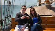 A man and a woman are smiling and toasting with drinks aboard a sailboat on a sunny day.