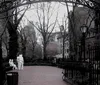A grayscale image showing a serene park pathway with benches leafless trees a street lamp and sculptures depicting people in an urban setting