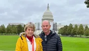 Two people are standing in front of the United States Capitol building, which appears to be partially under scaffolding.