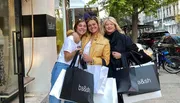 Three people are smiling for the camera on a city sidewalk, holding multiple shopping bags from a store named 