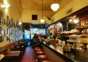 The image depicts the cozy interior of a classic diner with patrons seated at the counter and framed pictures lining the wall.