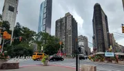 The image captures a bustling city street corner with diverse architecture, from modern skyscrapers to classic buildings, under a cloudy sky.