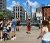 People are enjoying a sunny day on an urban walkway with modern buildings and skyscrapers in the background