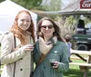Two smiling women are holding drinks at an outdoor event with tents and a sign reading Grand Open Osprey in the background