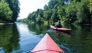 Two people are kayaking on a calm river surrounded by lush greenery.