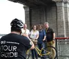 A group of smiling people wearing helmets are standing with bicycles on a sunny boardwalk by the water with a bridge in the background