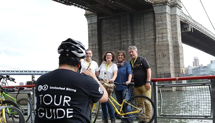 A tour guide wearing a helmet and a shirt labeled TOUR GUIDE is taking a photo of a group of four cheerful people with bicycles by a bridge.