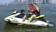Two people are making peace signs while riding a Yamaha WaveRunner on the water with a city skyline in the background.