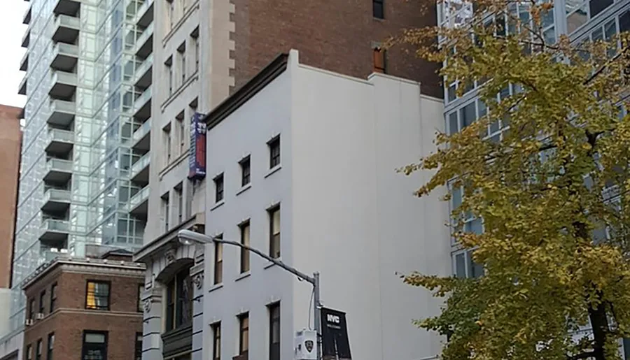 The image shows a streetscape with a mix of architectural styles including a modern glass building, older brick buildings, and a white facade without windows, under an overcast sky with some autumn foliage visible.