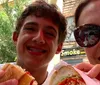 Two smiling people are taking a selfie while enjoying sandwiches outdoors