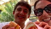 Two smiling people are taking a selfie while enjoying sandwiches outdoors.