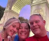 Three people are taking a smiling selfie in front of an illuminated arch with a twilight sky in the background