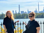 Two smiling people pose in front of a waterscape with a city skyline in the background on a sunny day.