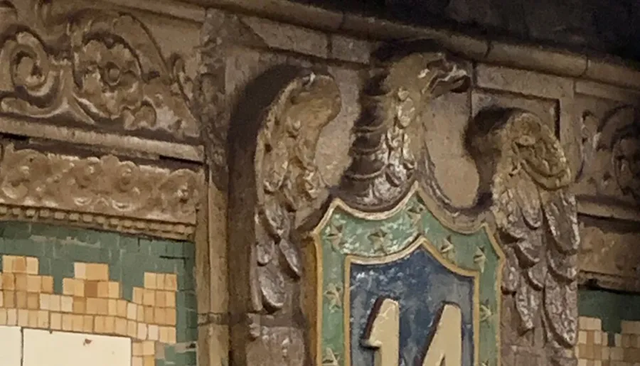 The image features an ornate architectural detail with sculpted figures flanking a decorative shield bearing the number 14, set against a wall with vintage tilework.