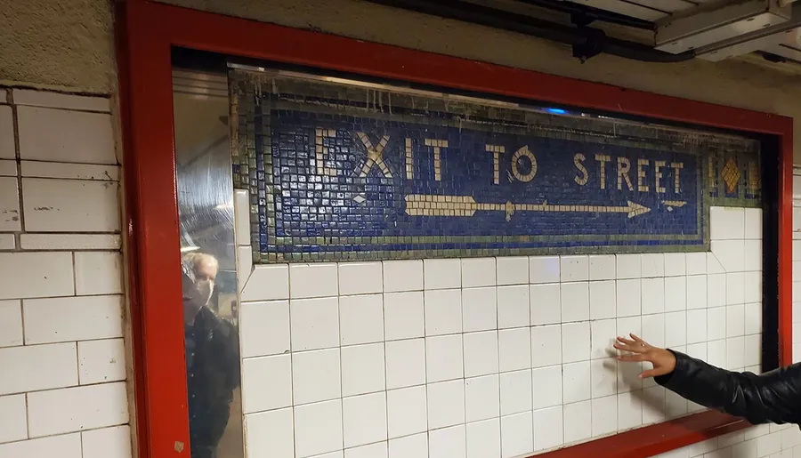 The image shows a vintage mosaic tile sign reading EXIT TO STREET on a subway station wall, with a reflection of a person visible in the glass and another person's hand pointing.