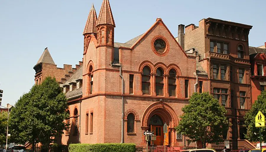 This image features a Victorian Gothic-style red brick building with pointed arches and a turret, set among other older buildings on a sunny day with clear skies.