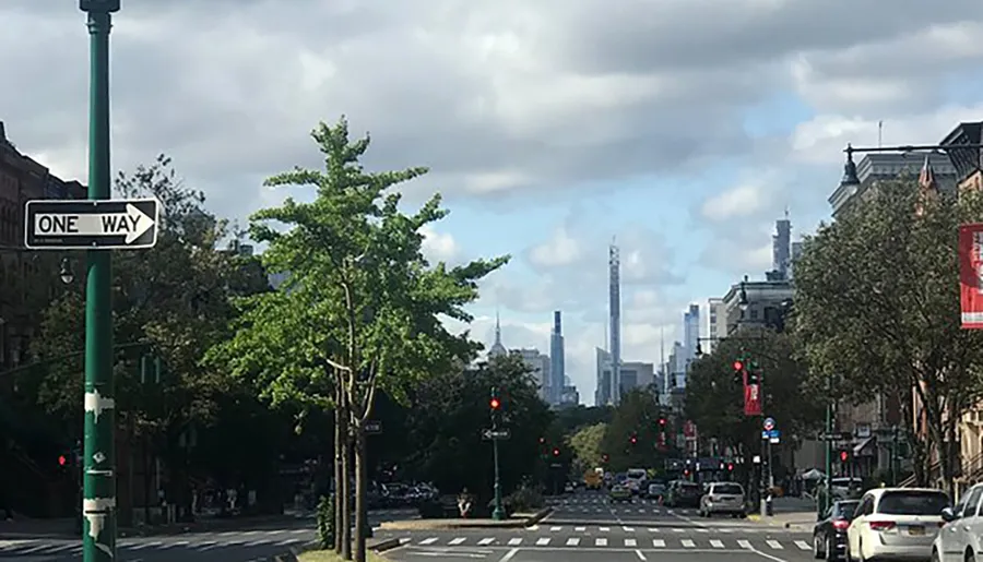 The image captures a city street view with cars, traffic lights, an overhead 'One Way' sign, with a backdrop of a distant modern skyline under a cloudy sky.