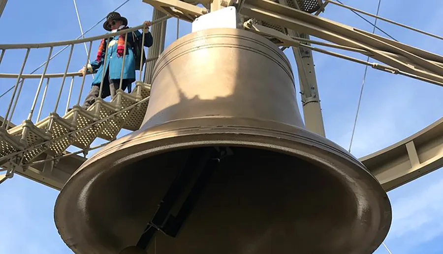 A person in a traditional outfit stands near a large bell suspended within a metal framework against a clear sky.