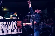 A passionate performer is raising his index finger to the sky while singing into a microphone at a lively piano bar, with an audience enjoying the show in the background.