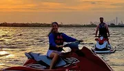 Two people are riding jet skis on a body of water with a sunset in the background, and a city skyline is faintly visible in the distance.