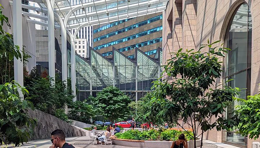 The image shows a modern urban atrium with a glass ceiling, surrounded by buildings, and featuring lush greenery and seating areas where people can relax.