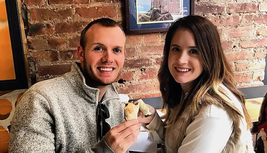 Two people are smiling at the camera while holding food, sitting in a cozy eatery with a brick wall in the background.