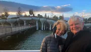 A smiling couple poses for a photo in front of a unique bridge over water at dusk.