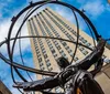 A statue of Atlas holding the celestial spheres stands before the art deco facade of a skyscraper under a blue sky with scattered clouds