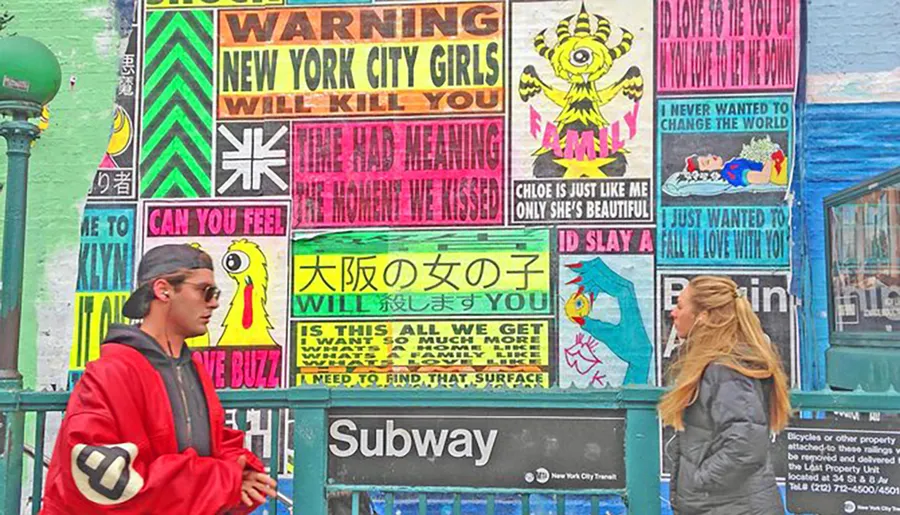 Two people are standing in front of a colorful wall filled with vibrant posters and urban art near a subway entrance.