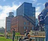 A person is taking a photograph while others enjoy a sunny day on a rooftop garden overlooking a mix of historic and modern urban architecture
