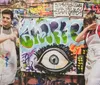 Two people are joyfully displaying their graffiti artwork featuring a large eye with a dynamic and colorful backdrop full of street art