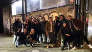 A group of smiling people is posing for a photo on a city street at night, with graffiti in the background.
