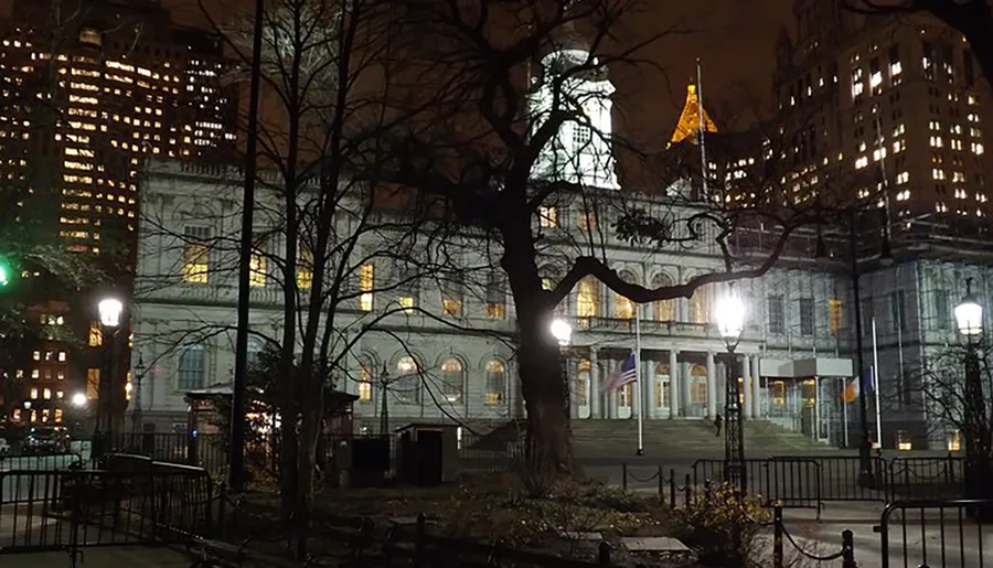 An illuminated historic building stands prominently at night amidst the silhouettes of bare trees and the backdrop of a modern cityscape.