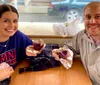 A smiling man and woman likely in a caf or bakery are holding up half-eaten chocolate muffins with the woman wearing an Alumni sweater and the man wearing a light jacket