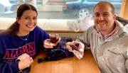 A smiling man and woman, likely in a café or bakery, are holding up half-eaten chocolate muffins, with the woman wearing an 